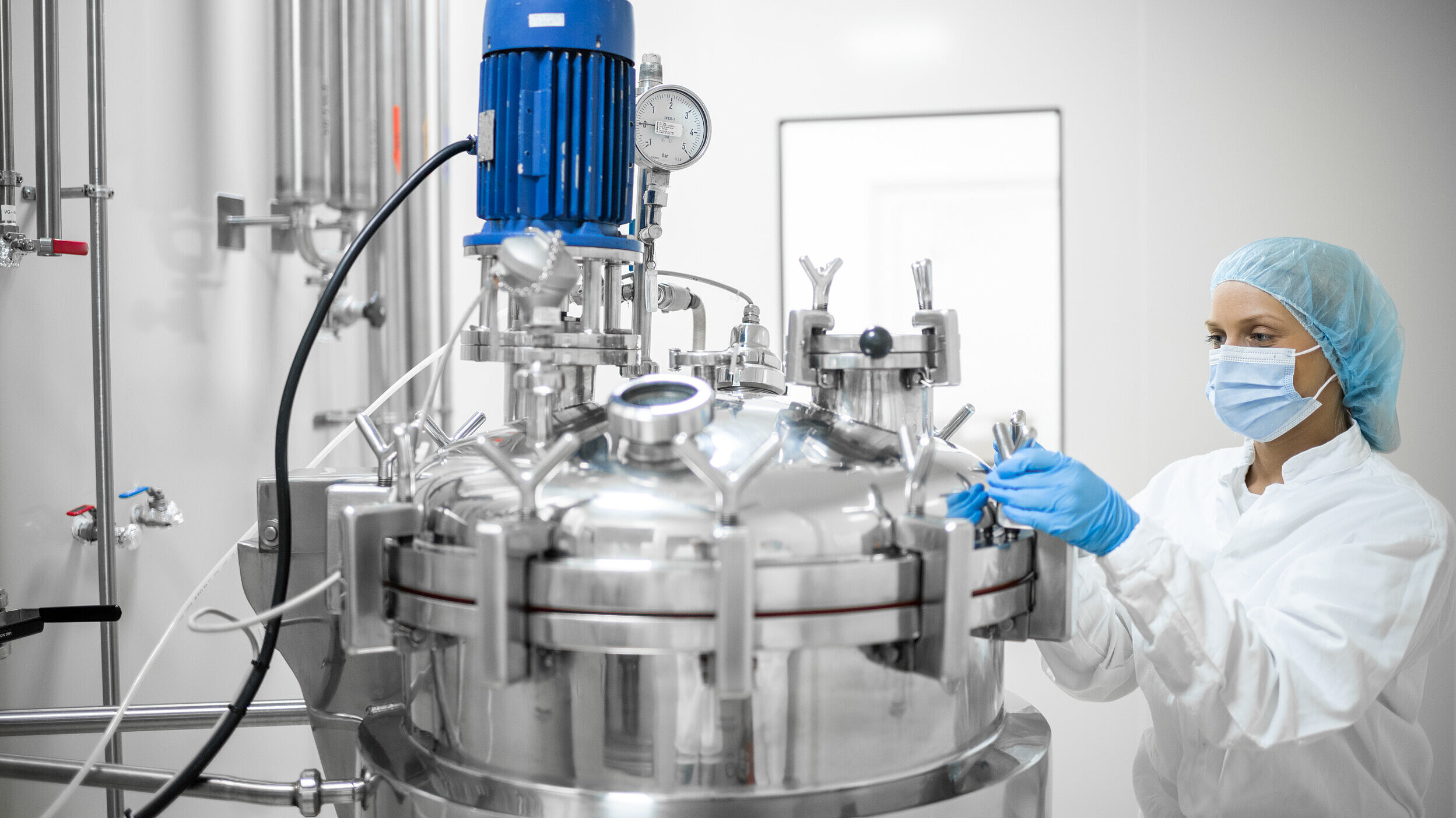 „Mitarbeiterin in Schutzkleidung arbeitet an einem modernen Edelstahltank in einem sterilen Labor oder Pharmaunternehmen – Biotechnologie, pharmazeutische Produktion, Reinraumtechnik